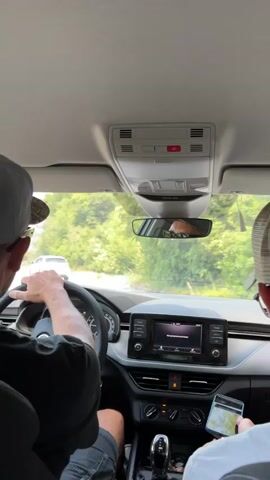 The video is filmed from inside a car, looking forward. The driver, wearing a hat, has their hands on the steering wheel, and is looking ahead. To the right, a passenger wearing a baseball cap is holding a smartphone. The car's dashboard is visible, with a touchscreen infotainment system displaying a song title and artist. The car is on a paved road, with green foliage and trees on either side. A white car is visible ahead on the road, moving in the same direction. The sky is clear and bright. The video captures a moment of travel.