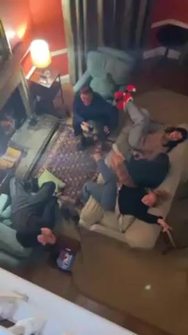 From a high vantage point, a living room is shown. A man sits in a chair, holding up one finger. On a large sofa, four people are laying down, with their feet elevated. A fireplace is to the left.