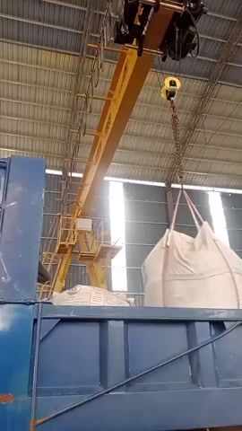 A yellow overhead crane is lifting a large white bulk bag, suspended by straps and a chain, over the blue bed of a truck. The bag is partially over the truck bed, and a person wearing an orange vest is visible in the truck bed, looking up at the bag. The scene takes place inside a large building with a corrugated metal roof and walls.