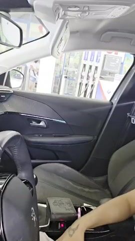 A young person wearing sunglasses and a striped shirt sits in the passenger seat of a car, making a pouting face at the camera. The car appears to be at a gas station, with gas pumps visible through the window. Another person, wearing a white shirt, is visible outside the car.