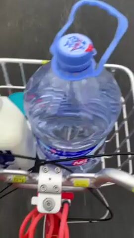 The video shows a person riding a bicycle with items in the front basket. The items include a large bottle of water, a bottle of cleaning liquid, and possibly a smaller container. The rider is on a paved path, likely a bike lane, adjacent to a highway. The weather is overcast and wet, with puddles visible on the path. In the background, there is a grassy area with trees and a decorative stone railing. The landscape includes a tree-covered hill. The camera is mounted on the bike, providing a first-person view of the ride.