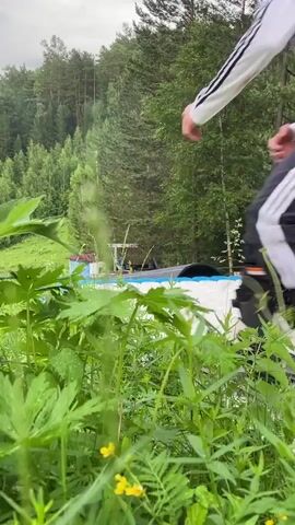 The video shows a person on a snowboard in mid-air over a white surface, possibly an inflatable landing pad, with a forest in the background. The person is wearing a helmet and protective gear, including a white and black jacket with grey patterns, and black pants with orange padding. The person is captured in motion as they perform a jump. Lush green vegetation is in the foreground, with some small yellow flowers visible among the leaves. The sky appears overcast. The scene suggests an outdoor recreational activity, possibly a ski or snowboard park.