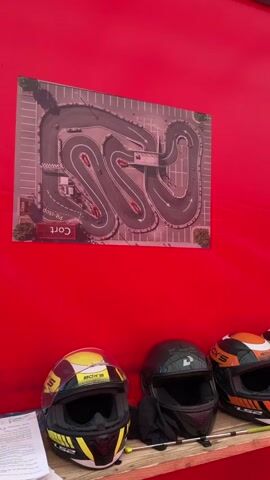 The video shows a close-up of three helmets lined up on a shelf. Behind the helmets, a red fabric wall is visible. Above the helmets, a poster of a go-kart track is mounted on the wall. The helmets are of various colors, with the leftmost one being yellow and black, the middle one black, and the rightmost one orange and black. All three helmets appear to be full-face motorcycle or go-kart helmets. There is a stack of papers on the left side of the shelf, partially obscured by the yellow and black helmet. The lighting is bright, and the scene appears to be outdoors or in a well-lit indoor space.
