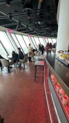 The video shows the interior of a high-altitude bar or restaurant with large windows providing a panoramic view of a city. Several people are seated at tables along the windows and standing near the bar area.  The bar features a countertop and display area with cups. The floor is red with speckled pattern. The overall lighting is a bit dim, with artificial lights in the ceiling and a touch of pink lighting near the windows. The outdoor scene reveals the urban sprawl of a city on an overcast day. There are many buildings, roads, and a highway interchange visible below.