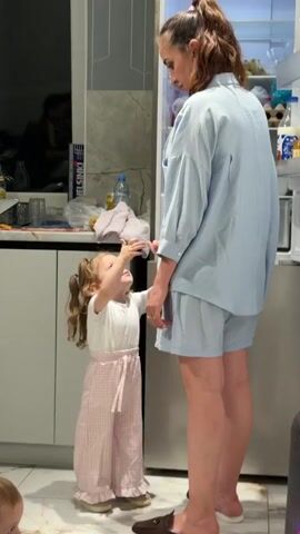 A woman and a small child are standing in a kitchen. The woman is wearing a blue shirt and shorts, and she has her hair up in a ponytail. The child is wearing a white shirt and pink plaid pants. The woman picks up the child and holds her close in front of the open refrigerator. Another child is partially visible on the bottom left. There are various items visible inside the refrigerator. The kitchen has grey cabinets and a tiled backsplash. There are some items on the counter near the sink, including a box of crackers. Some music is playing.