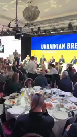 The video depicts a large, formal gathering taking place at what appears to be a conference or summit, possibly related to Ukraine. A large screen displays the words "UKRAINIAN B" and "Davos, Switzerland" in white text against a blue and yellow background reminiscent of the Ukrainian flag. Numerous people, mostly men in suits, are seated around large, round tables set with plates, glasses, water bottles, and unfinished food. Servers in white shirts move between the tables, offering food on trays. Some attendees are talking among themselves, while others seem to be listening to a speaker. In the foreground, the back of someone's head with a distinctive shaved hairstyle is visible. The lighting in the room is bright, with spotlights directed at the stage area. The overall atmosphere is professional and formal.