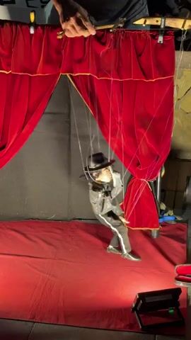 A puppet show is taking place outdoors at night. A single puppet dressed in a black suit, white shirt, and black hat is center stage, suspended by strings from above. The puppet is performing an action resembling a dance or fight move. To the left, another puppet dressed in a yellow suit and red cape is visible, also suspended by strings. The stage is set with red velvet curtains and a red carpet. Lighting equipment is visible at the bottom of the frame. A person's hands are visible at the top of the frame, manipulating the puppet's strings.