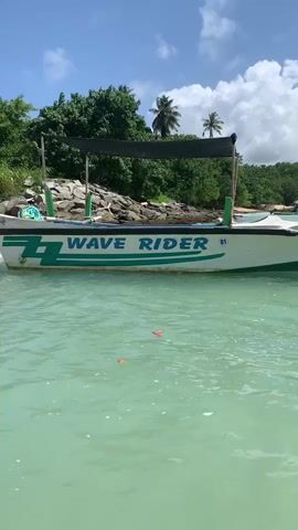 This is a low-angle, medium shot of a white boat with green trim that reads "WAVE RIDER" in teal lettering. The boat is in calm, turquoise water. Behind the boat, a sandy beach is visible with large rocks and lush green trees and palm trees. The sky is blue with scattered white clouds. There are two orange objects floating in the water in front of the boat. The boat appears to be stationary.