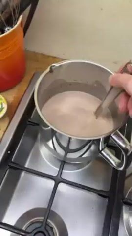 Here's a detailed description of the video:

The video shows a person stirring a light-brown liquid in a stainless steel saucepan on a gas stovetop. The liquid appears to be simmering, with small bubbles visible on the surface. The person is using a metal whisk to stir the liquid thoroughly. The stovetop is stainless steel with black burner grates. To the left of the saucepan, there's an orange ceramic utensil holder with various kitchen utensils inside. There's also a small, shallow dish with a green and yellow pattern on the wooden countertop. The focus is on the liquid being stirred in the saucepan. The person's hand and arm are visible while stirring.