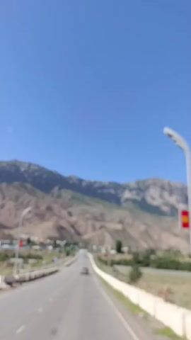 THE VIDEO SHOWS A DRIVING VIEW OF A ROAD WITH MOUNTAINS IN THE BACKGROUND. A CAR IS VISIBLE ON THE ROAD AHEAD. THERE ARE STREET LIGHTS AND GUARDRAILS ALONG THE SIDE OF THE ROAD. THE SKY IS CLEAR AND BLUE. THERE IS SOME VEGETATION ON THE SIDES OF THE ROAD AND IN THE DISTANCE. THE SCENE APPEARS TO BE IN A RURAL OR SEMI-RURAL AREA.