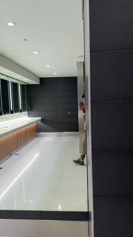 Here's a description of the video:

A man in a beige hoodie and sweatpants takes a selfie in a modern bathroom. He is wearing a black fanny pack and backpack. The bathroom has a long row of sinks and mirrors on one side, and gray tiled walls. The floors are white tile. The man has short brown hair and is wearing a watch. He appears to be alone in the bathroom.