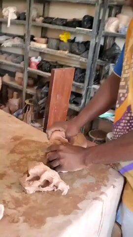 Here is a description of the video:

The video shows a person working with clay on a table. The person uses their hands to mold and shape the clay. There's an object on the table which looks like a skull in its early stages. The table surface is stained with clay residue. In the background, there is a shelving unit filled with various clay objects.