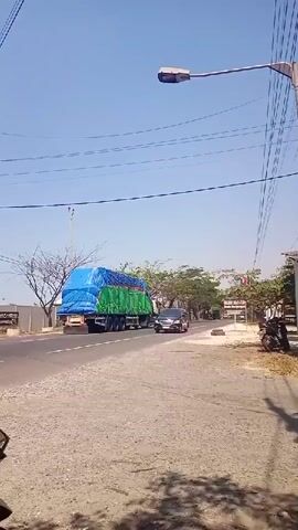 This video shows a street scene in Sumurtawang, Indonesia, on a bright, sunny day. The sky is a clear blue, suggesting fair weather. The main focus is a black SUV driving on a paved road. The SUV appears to be an Innova model, judging by the visible badging on the front. Its license plate reads "B 1126 UOH."

Behind the SUV, a large truck is also traveling down the road. The truck is green and has a large, bright blue tarp covering its cargo. The tarp is pulled taut, indicating the cargo is likely being protected from the elements. The truck is considerably larger than the SUV, suggesting it is carrying a significant load.

The road itself is asphalt, with a gravel shoulder on the right side of the frame. There are power lines strung overhead, supported by poles. A street lamp is visible on the right. On the left side of the road, there is a fence and some sparse, leafless trees, characteristic of a dry season or certain types of vegetation. On the right side, there are more trees with green foliage, and a motorcycle is parked.

The overall impression is one of a typical day on a road in a possibly rural or semi-urban area of Indonesia. The bright sunlight and clear sky suggest it is daytime, likely mid-morning or afternoon. There are no people prominently visible, but the presence of vehicles indicates ongoing activity. The scene is peaceful and ordinary, with no apparent signs of distress or urgency. The visible text is limited to the license plate of the SUV.