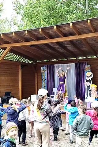 A woman in a purple dress and gold cape is on a wooden stage, dancing in front of a group of children. The children are moving their arms, some with hands raised in the air, seemingly imitating the woman. Behind her is a banner that says “Welcome” and another standee of a character in black. The stage is under a wooden roof. The setting appears to be outdoors, surrounded by trees.
