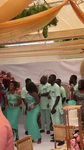 Sure, here is a description of the video:

The video shows a group of people dancing at an event, possibly a wedding. The people are dressed in colorful clothing, mostly in mint green, and appear to be enjoying themselves. There is a banner in the background with the names of the people being celebrated: "OLUWON AND EMMANUEL." There's a tiered wedding cake near the right corner. There is a tent with beige and orange draping overhead. A person in a white dress, with what appear to be diamond-like embellishments, moves away from the camera. Another person walks by in the background with a white shirt covered in a black, white, and brown design.