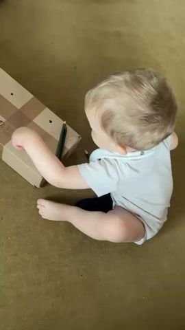 A baby is sitting on a brown carpet in front of a cardboard box. The baby is wearing a light-colored onesie and dark-colored socks. The baby is reaching out to a green pencil that is resting on the cardboard box. The baby's left hand is holding the pencil and the baby appears to be drawing on the box. There is another pencil on the carpet to the right of the box. The baby's right hand is also on the carpet.