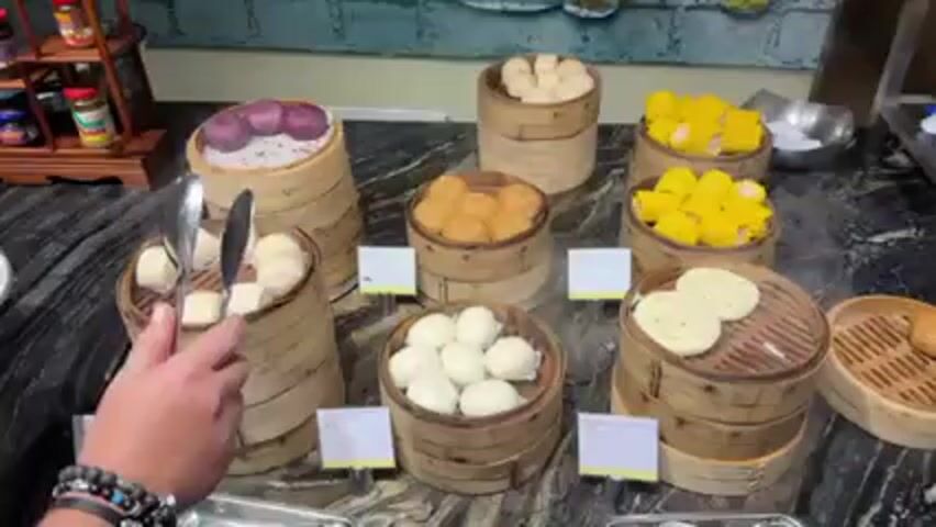 A person is using tongs to pick up purple steamed buns from a bamboo steamer at a buffet. There are several other bamboo steamers on a black marble counter, each containing a different type of steamed bun or food. The other steamers contain plain white buns, golden buns, yellow buns, and white disk-shaped buns. The person is wearing a white shirt and beaded bracelets. There are small cards in front of each steamer, presumably labeling the food. In the background, there are condiments and wall art. Steam is visible rising from the steamers, indicating the food is hot.
