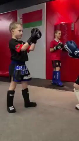 The video shows four young boys, wearing boxing gloves, sparring in a boxing gym. The gym has grey mats, with a red and white padded wall that has a small flag on it. One of the boys is wearing blue and black shorts and the others have black shorts. The boys are actively moving around, throwing punches, and blocking. There is a boxing ring in the background. A blue punching bag hangs from the ceiling.
