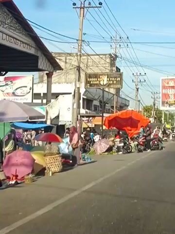 The video shows a busy street scene, likely a market, with several stalls and vendors. Numerous people are present, some on foot and others on motorcycles. There are a variety of goods on display, including what appear to be fruits and vegetables, as well as textiles. Several motorcycles are parked along the side of the road, and some individuals are seen riding them. The environment is outdoors, with buildings and power lines visible in the background. The overall impression is one of a vibrant and active marketplace.
