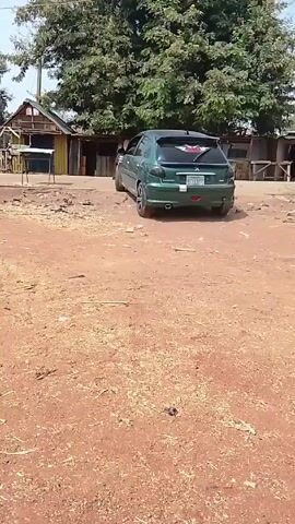 The video shows a green car backing up and driving away on a dirt road. There are several small, wooden buildings and a large tree in the background. The ground is dry and covered in loose debris. The sky is visible above the tree.