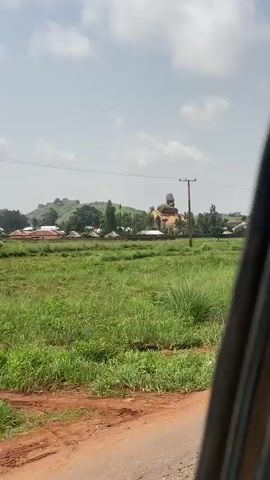 This is a video taken from a moving vehicle, likely in a rural area of Hoss, Nigeria, during the daytime. The scene is dominated by a vast expanse of tall, green grass, with a dirt path visible in the foreground. In the middle ground, there are several small, thatched-roof huts and some modern buildings with corrugated metal roofs, suggesting a small village. A prominent feature in the background is a large rock formation on a hillside, with a distinctive shape that resembles a seated figure. Several trees are scattered throughout the landscape, and a wooden utility pole with wires runs across the frame, indicating the presence of electricity. The sky is partly cloudy, suggesting fair weather. The overall impression is one of a peaceful, rural landscape with a unique natural landmark. There is no visible text in the video.