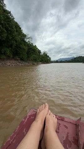 The video shows a first-person perspective of a person's bare feet resting on the maroon surface of a boat. The boat is moving on a wide, muddy brown river. On the left side of the river, a dense, green forest covers a sloping embankment. The sky is overcast with dramatic, gray clouds. In the distance, the river curves, and mountains can be seen. The water is choppy with visible ripples.