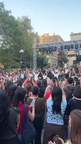 The video shows a large crowd of people gathered outdoors, facing a stage where performers in white outfits are dancing. The crowd is clapping their hands and many are holding up their phones, presumably to record or take pictures. The setting appears to be a public square or street, with a building visible behind the crowd and trees in the background. The lighting suggests it is daytime, possibly late afternoon or early evening. The overall atmosphere is festive and celebratory.