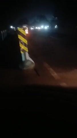 The video is a blurry, dark recording of a road at night. In the foreground, a yellow and black striped traffic bollard is visible on the left. Further down the road, there are multiple vehicles with their headlights on, creating bright streaks of light. One vehicle, possibly a truck, has orange lights illuminated. The overall impression is of a dimly lit highway with traffic.