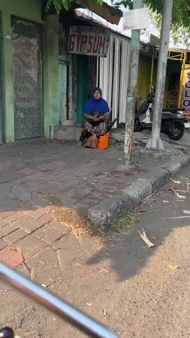 A woman in a blue headscarf and a patterned dress sits on a curb next to a motorcycle. She is holding a dark object, possibly a phone, and there is luggage beside her. The scene appears to be outdoors, with buildings and trees in the background. There is debris on the ground, including trash and what looks like spilled liquid.