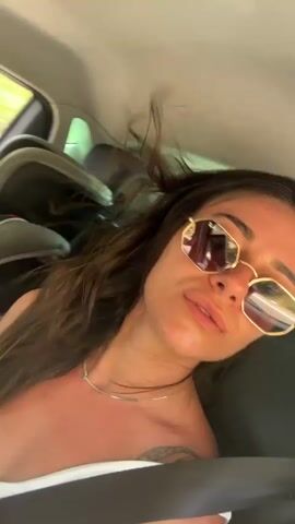 The video shows a woman in the back seat of a car. She is wearing sunglasses and a white top. She is smiling and touching her hair. The car appears to be moving. There are child car seats in the background. The lighting suggests it is daytime and the weather is sunny.
