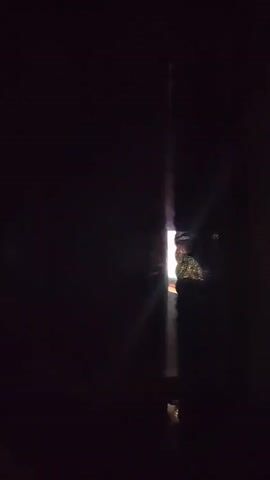 The video shows a dark room with a small opening providing a glimpse into another area. A bright light shines through the opening, illuminating a portion of the dark room. The source of the light appears to be a fire, and the opening is narrow, creating a vertical beam of light in the dark space. There is some texture and a reflective surface visible near the opening.
