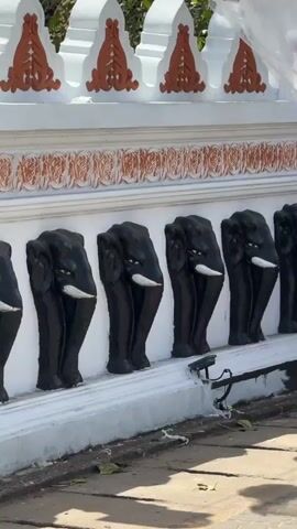 The video showcases architectural and sculptural details of a Buddhist temple in Matara, Sri Lanka, during the daytime under bright sunlight.

The initial view focuses on a white wall adorned with repetitive black elephant statues, each featuring white tusks, protruding from its surface. Above the elephants, the wall features an ornate orange and white decorative pattern.

As the camera pans left, a colorful Buddhist flag, with stripes of red, yellow, blue, white, and orange, becomes visible on the left side of the frame, gently swaying. Below the wall, a carved stone lion statue is situated, and a grey mailbox is attached to an adjacent white pillar.

The pan continues upwards, revealing a large, grey stone Buddha statue seated on a platform. Below the statue, a grand set of dark grey stone stairs with intricate carvings on the risers and along the banisters ascends. The surrounding area features lush green trees and other temple structures in the background, all under a clear blue sky. A plaque is visible near the Buddha statue, but the text is unreadable. No people are visible in the video.