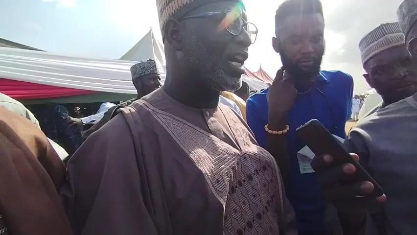 The video captures a close-up shot of a gathering of people, seemingly outdoors under a bright, slightly overcast sky. The central figure is a middle-aged man with graying hair and a beard, wearing glasses and a traditional brown robe with intricate patterns, topped with a striped, woven cap. He appears to be speaking. Surrounding him are other men, some in traditional attire including caps, and one in a casual blue shirt holding a smartphone. In the background, there's a glimpse of tents with white and red fabric, suggesting the event is set up for a large number of attendees. The camera angle is relatively low, looking up at the primary figure as he addresses the crowd.