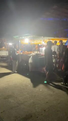 The video shows a night scene at an outdoor market or fair. Several food stalls are visible, lit by bright lights. People are milling around, some standing in lines at the food stalls, others walking through the area. The stalls appear to be selling various types of food and drinks. The ground is paved and there are some trees or buildings in the background. The scene appears lively with people interacting.
