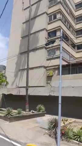 This video shows a moving perspective, likely from a vehicle, traveling along a street in Macuto, Venezuela. The scene is dominated by a tall, beige apartment building with many windows and balconies. The building appears to be older, with some signs of wear. In front of the building, there is a landscaped area with various plants, including agave-like plants and some flowering bushes.

Several trees line the street, casting dappled shadows on the asphalt. The perspective shifts as the vehicle moves, revealing more of the building's facade and the surrounding greenery. In the foreground, the yellow and white lines of the road are visible. A utility pole with wires is also present.

As the video progresses, the camera passes by a small collection of flags, including one that appears to be the Spanish flag, suggesting a possible event or establishment with international ties. Further down the street, a motorcycle is parked near a tree. The overall impression is of a sunny day in a residential or mixed-use urban area, possibly with a tropical or subtropical climate given the vegetation. The light suggests it is daytime, likely morning or afternoon, with clear skies. There are no prominent emotional cues, but the scene is calm and ordinary. No discernible text is visible in the video.