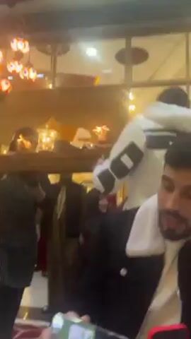 Inside a restaurant, two men are carrying a large tray filled with food and drink across the room. The tray appears to be quite heavy, as both men are using considerable effort to support it. A man is looking at his phone. Seated at tables are several customers who are watching the procession. The restaurant is dimly lit. The camera is moving throughout the room, tracking the tray being carried.