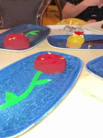 The video displays a table at what appears to be a restaurant, where four decorative desserts are presented on rustic-style blue plates. Each dessert is unique in its appearance and design: one is a red rose with gold flecks and a green stem, another resembles a yellow tiered cake with a red rose embellishment, another is a small grey cake shaped like a tea cup, and the last is a tart topped with white cream and a decorative disc. In the background, a person with a polka dot shirt and a yellow hat can be seen. The table is light colored and partially reflects the light. The camera slowly pans over the arrangement of desserts.