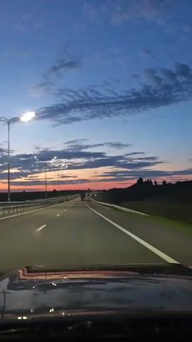 The video shows a first-person view from a vehicle driving on a highway at dusk. The sky is a gradient of deep blue at the top, transitioning to lighter blues and pinks near the horizon, with scattered clouds. Streetlights are visible along the side of the highway, and their lights are illuminated. The highway has multiple lanes, indicated by white lines, and a guardrail separates the lanes from the oncoming traffic. In the distance, the lights of other vehicles can be seen. The road surface appears to be asphalt. Trees and vegetation line the sides of the highway. The dashboard and windshield of the vehicle are visible in the foreground, reflecting the lights and the sky.