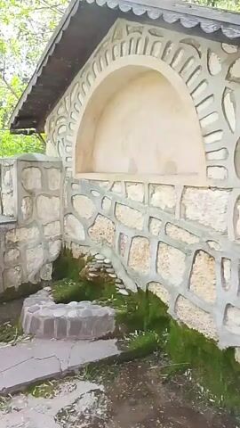 The video shows an outdoor scene featuring a stone structure with an arched niche. In front of the structure is a stone path leading to stone stairs that ascend a small hill covered in greenery and flowering trees. A small wooden structure sits at the top of the hill.  The video appears to be taken on a sunny day.  There are no people visible in the video.