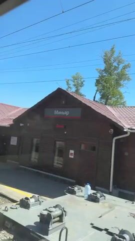The video was filmed from a train window while the train was moving at slow speed, perhaps approaching or departing a station. The sky is clear and bright blue. The landscape is rural, including trees and grassy areas. In the first few frames, a dark brown building is visible. Then a white building. Finally, a few houses and cars are visible through the train window. Power lines are strung up in the sky throughout the video.