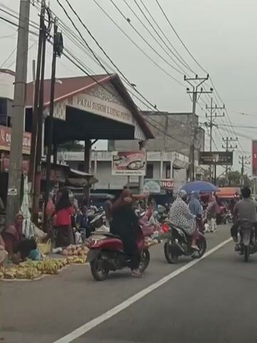 The video shows a street scene in what appears to be a market area. There are several motorcycles and people on and around them. Some people are dressed in traditional Islamic clothing, including headscarves and long robes. In the background, there is a building with a sign that reads "PASAI SUBOH LUENG PUTU". There are also market stalls set up with goods, likely produce, displayed on the ground. The sky is overcast, and there are numerous power lines visible overhead. The overall atmosphere suggests a busy marketplace.