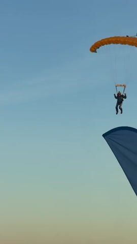 A skydiver descends from the sky with a yellow and blue parachute. The skydiver is wearing a blue jumpsuit and a green helmet with a camera attached. The skydiver is approaching a grassy field with trees in the background. A blue and white banner with text is visible in the foreground, partially obscuring the view of the skydiver. The sky is clear and blue, with a hint of sunset colors. The skydiver is in the process of landing.