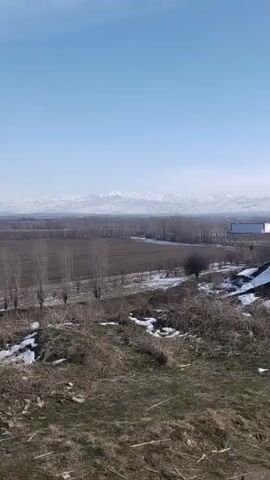 The video shows a landscape with a view of snow-capped mountains in the distance under a clear, light blue sky. In the foreground, there's a hillside covered with dry grass and some patches of snow. The middle ground consists of a flat, agricultural area with bare fields, rows of leafless trees, and a couple of buildings. The overall scene appears to be in a rural or semi-rural location during the late winter or early spring.