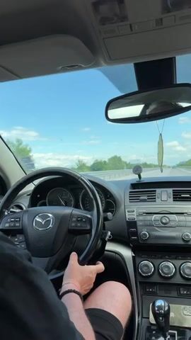 The video shows a driver in a black Mazda, likely on a highway. The driver is wearing a black shirt and shorts, with a bracelet on their left wrist. The car's dashboard is visible, including the steering wheel, instrument panel, and center console. A leaf-shaped air freshener hangs from the rearview mirror. The sky is blue with some clouds, and trees and billboards line the side of the road. Other vehicles can be seen in the distance. Music is playing.