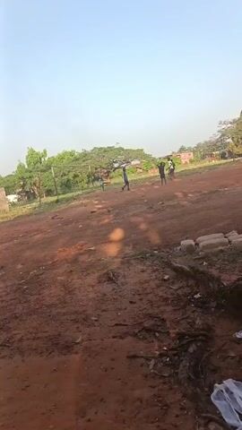The video shows a group of boys playing soccer on a dirt field. The field is surrounded by trees and some buildings are visible in the background. In the foreground, there is a large tree root and some trash. The sky is clear and blue. The boys are running and kicking the ball, seemingly enjoying their game.