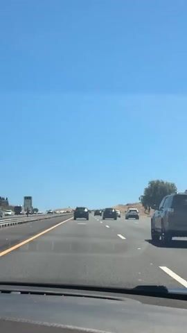 The video shows a view from the driver's seat of a car traveling on a multi-lane highway. The sky is clear and blue. There are several cars on the highway ahead, traveling in the same direction. The car in front of the viewer is a dark colored SUV. On the left side of the highway, there is a guardrail and some signs. The landscape on the right side of the highway is hilly and dry, with some trees. The video appears to be taken during the daytime.