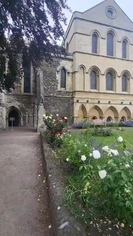 The video shows a slow pan across a garden area adjacent to a large stone building. The building has arched windows and appears to be old, with some sections in ruin. In the foreground, there are flower beds with roses and lavender. A pathway is visible on the left side of the frame. The sky is overcast.