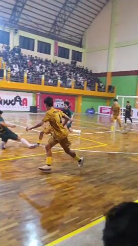 A futsal match is in progress indoors on a wooden court. Spectators fill the stands on the left and right sides of the court. In the foreground, a referee in a pink shirt and black shorts faces away from the camera, observing the game. On the court, players in yellow and green uniforms are actively engaged in the match. One player in a yellow jersey is on the ground near the center of the court, possibly having fallen. Another player in green is positioned near him. The ball is visible on the court. In the background, banners and advertisements are displayed on the walls of the indoor arena. The lighting appears to be artificial, originating from overhead fixtures, casting reflections on the polished court surface.