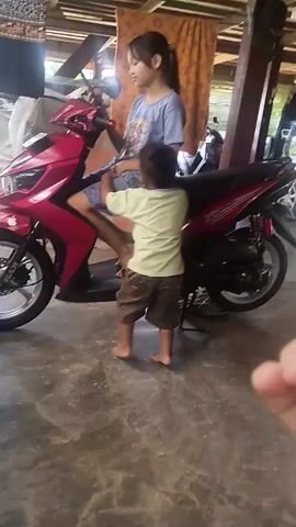 A young girl is sitting on a red motorcycle. A young boy stands in front of her, facing the camera, and he is reaching for the girl. The girl appears to be handing the boy something. The background consists of a wooden structure and a concrete floor. There is another motorcycle and a black vehicle visible further in the background. The lighting is somewhat dim, with a strong beam of light coming from the upper left. The perspective of the video appears to be from someone standing in front of the children.