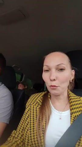 A woman with fair skin and long blonde hair sits in the passenger seat of a car, filming herself and others with a smartphone. She is wearing a white shirt and a yellow houndstooth jacket, and she has a pendant necklace. The man driving the car is fair-skinned, with dark short hair and a gray t-shirt. There are at least two other people in the back seat of the car, one of whom is a child wearing a baseball cap. The woman is smiling and looking at the camera, and the man is looking straight ahead as he drives. The car is moving, and trees are visible outside the window. Loud music is playing. The woman is wearing a seatbelt.
